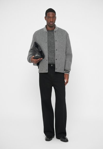 Γκρι ζακέτα από μάλλινη σύνθεση με ριμπ μανσέτες, μαύρο πλεκτό πουλόβερ, και μαύρο παντελόνι σε φαρδιά γραμμή. Κρατά μια μαύρη δερμάτινη τσάντα στο αριστερό χέρι.
