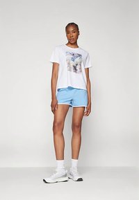 Wit T-shirt met korte mouwen en een kleurrijke afbeelding op de voorkant, gecombineerd met blauwe korte broek en witte sportschoenen, staand op een neutrale achtergrond.