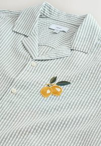 Jasnozielona i biała koszula na guziki w paski z haftowanymi żółtymi cytrynami i zielonymi liśćmi na piersi, krótki kołnierzyk, dwa białe guziki.