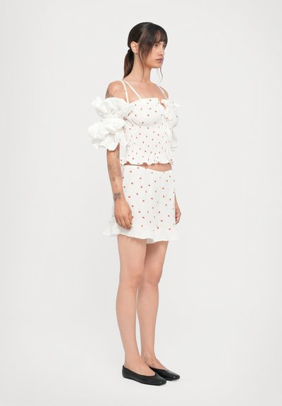 Hvid smock-top med røde prikker og pufærmer, parret med en matchende volang-miniskirt. Modellen har sorte flade sko på.