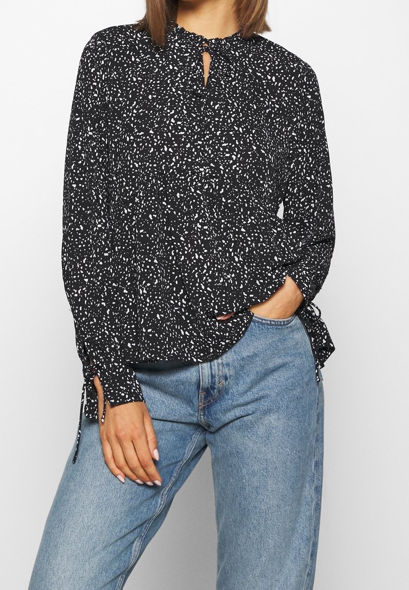 Blusa nera con motivo a pois bianchi, scollatura arricciata e lacci ai polsini. Abbinata a jeans di denim azzurro chiaro, con una forma rilassata.
