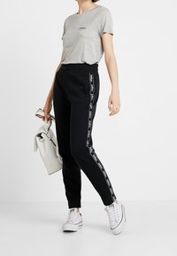 Šedé tričko s krátkým rukávem a logem, černé teplákové kalhoty s bílými bočními pruhy a bílé tenisky. Model drží bílou kabelku.