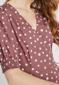 Frau in einem mauvefarbenen Kleid mit weißen Pünktchen, mit Puffärmeln und einem tiefen V-Ausschnitt, der einen Teil ihres Halses und ihrer Schulter zeigt.