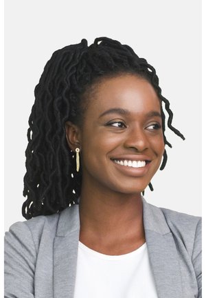 Kvinde med sorte dreadlocks iført en grå blazer over en hvid bluse. Hun har guld kædeøreringe, smilende mod en enkel baggrund.