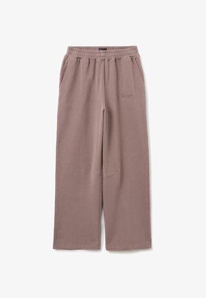 Pantalones de chándal de pierna ancha color marrón claro con cinturilla elástica, bolsillos laterales y un sutil texto bordado en la parte superior del muslo izquierdo.