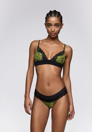 Vrouw met gevlochten haar, gekleed in groene en zwarte kanten lingerie met slangenprint, staand tegen een effen grijze achtergrond.