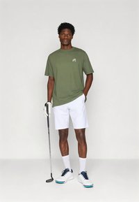 Bavlněné tričko olivově zelené barvy s krátkými rukávy, bílé šortky, bílé boty s modrými akcenty a golfová hůl v ruce. Na tričku je jednoduché logo.