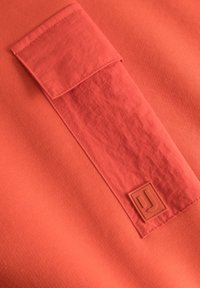 Koraal kleurige trui met een geptextureerd lichaam, een soepele zakflap en een rubberen logo-opdruk. Matte afwerking met contrasterende oppervlaktedetails.