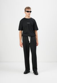 Tricou scurt cu mânecă neagră și logo, pantaloni plisati negri cu șnur alb, și ochelari de soare negri. Modelul stă în fața unui fundal alb.