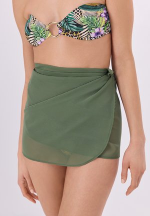 Žena má na sebe zelenú polopriesvitnú zavinovaciu sukňu zviazanú v páse a tropické vzorované bandeau bikinové tielko so zlatým krúžkom v detaile.