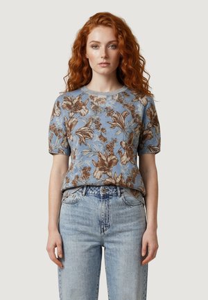 Kobieta o długich rudych włosach, ubrana w niebieską kwiecistą koszulę z krótkim rękawem, wpuszczoną w jasne, wysokowodne dżinsy, stojąca na tle jednolitego tła.