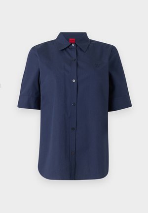 Marineblå kortermet skjorte med knapper, krage og rød etikett på innsiden av halsen, glatt front med subtil brodert logo på brystet.