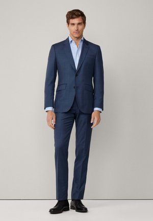 Dunkelblauer Herrenanzug mit einreihiger Jacke, Reverskragen und zwei Fronttaschen, kombiniert mit einem hellblauen Hemd und schwarzen Schuhen.
