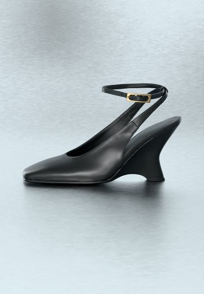 Μαύρο δερμάτινο παπούτσι slingback με χρυσή αγκράφα, τετράγωνη μύτη και κυρτή σφήνα τακουνιού σε ανοιχτό γκρι αντανακλαστική επιφάνεια.