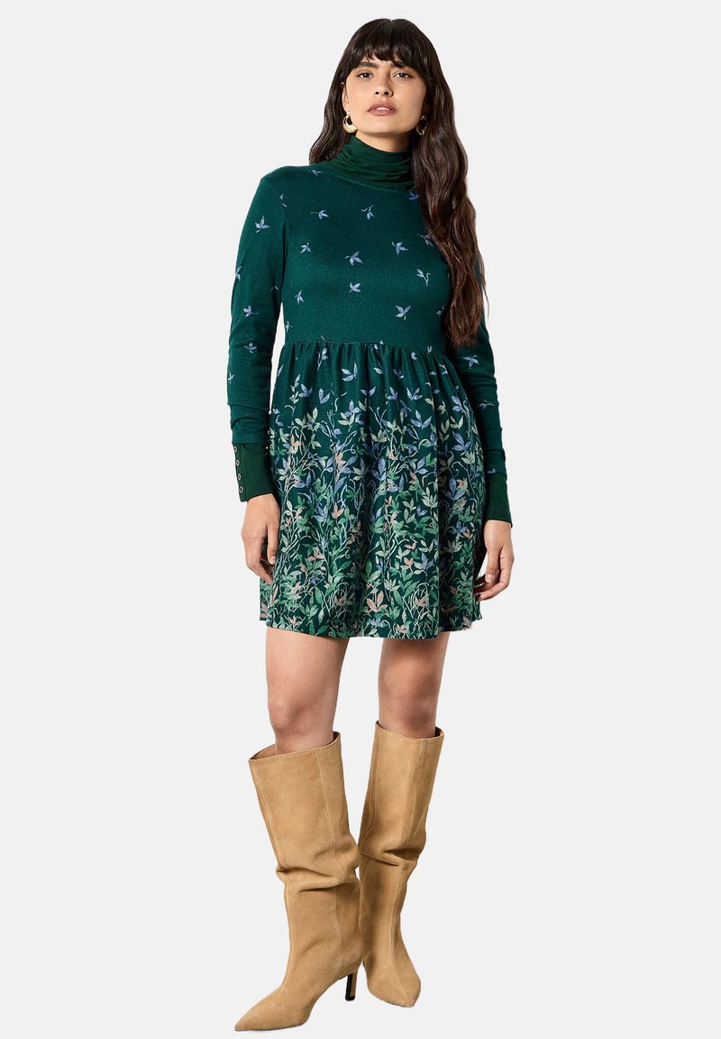 Abito verde con collo alto, caratterizzato da un motivo floreale sulla gonna. Indossato con stivali beige al ginocchio. Materiale morbido, corpetto aderente.