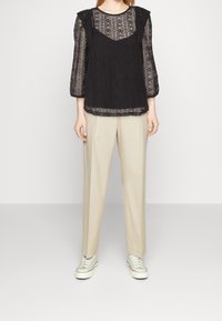 Top in pizzo nero con maniche lunghe a fantasia e scollo rotondo, abbinato a pantaloni beige su misura e sneaker verde chiaro.