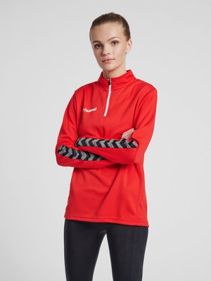 Ung kvinde med håret bundet tilbage bærer en rød sportspullover med sorte chevron-striber på ærmerne og sorte leggings, stående med armene over kors.