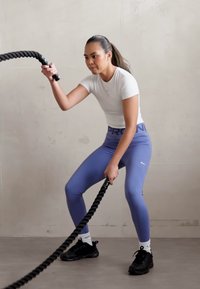 Sportaš u uskoj bijeloj majici i ljubičastim tajicama, drži debeli crni bojni konopac, stojeći na ravnom podu s neutralnom pozadinom zida.