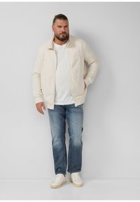 Homme avec une barbe portant une veste zippée crème, un t-shirt blanc, un jean bleu et des baskets blanches, se tenant contre un mur gris clair.