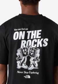 Osoba v černém tričku s bílým nápisem "Najdete mě NA SKALECH" a s karikaturou tancujících kostek ledu, plus logo The North Face.