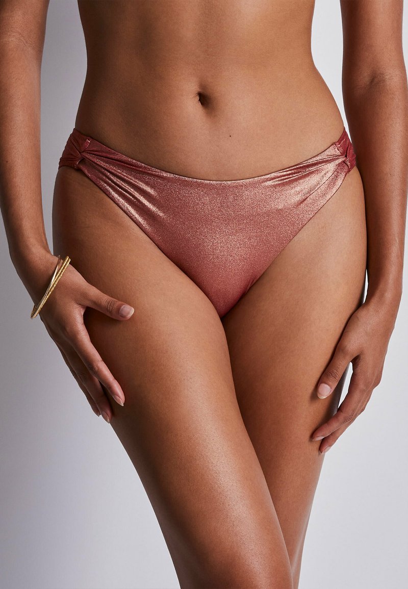 Femme portant un bas de bikini rose doré brillant avec un détail de nœud sur le côté et des bracelets dorés au poignet gauche, debout sur un fond uni.