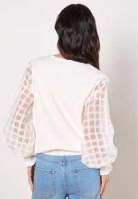 Weißer Pullover mit durchsichtigen, karierter Ärmelpartien und elastischen Bündchen, mit einem schlichten Rundhalsausschnitt und lockerer Passform. Kombiniert mit blauen Jeans.