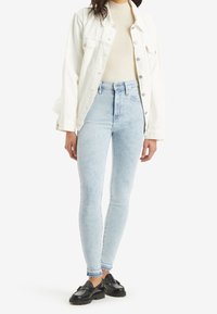 Jeans skinny de cintura alta en un azul claro con un lavado desvaído, combinados con una blusa de canalé en color crema y una chaqueta de denim blanca. Mocasines negros completan el look.