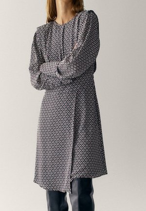 Frau trägt ein knielanges Kleid mit langen Ärmeln und einem schwarz-weißen geometrischen Muster, steht mit verschränkten Armen da.