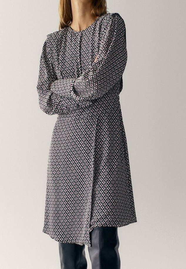 Femme portant une robe à manches longues, longueur genou, avec un motif géométrique noir et blanc, debout les bras croisés.
