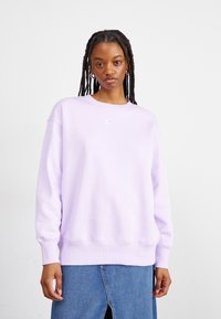 Jovem com cabelo trançado, usando brincos de argola prateados, um sweatshirt Nike lilás claro e uma saia de ganga azul com fenda frontal.