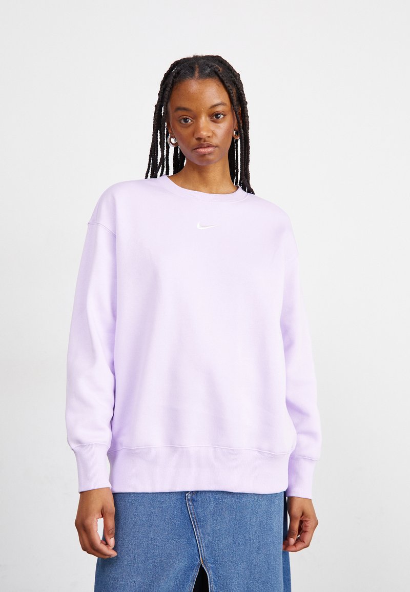 Jovem com cabelo trançado, usando brincos de argola prateados, um sweatshirt Nike lilás claro e uma saia de ganga azul com fenda frontal.