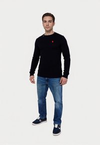 Mornarsko pleteno pulover s krožno odprtino in rdečim logotipom, v kombinaciji z modrimi jeans hlačami in temnimi supergami z belimi detajli.