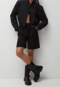 Schwarze, gekürzte Jacke mit Druckknöpfen, hoch taillierte schwarze Shorts und kniehohe schwarze Lederstiefel mit einer klobigen Sohle. Glatte Textur.