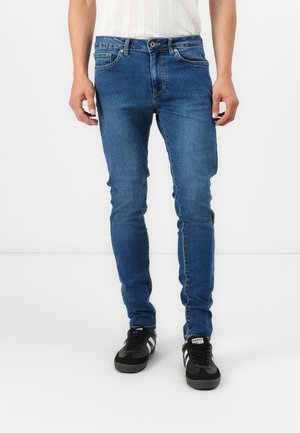 Personne portant un jean bleu slim et des baskets noires, debout devant un fond blanc uni.