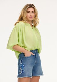 Blusa leggera verde chiaro e trasparente con chiusura a bottoni e vestibilità rilassata; abbinata a pantaloncini in denim blu a vita media con ricami floreali e orlo sfrangiato.