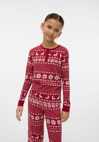 Dziewczynka w czerwono-białych piżamach na temat Bożego Narodzenia z wzorami z reniferami i płatkami śniegu, stojąca na tle czystego białego tła.