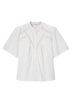 Jeune femme aux longs cheveux bruns portant un chemisier blanc texturé avec des détails en dentelle et un jean bleu clair, debout avec les mains sur les hanches.
