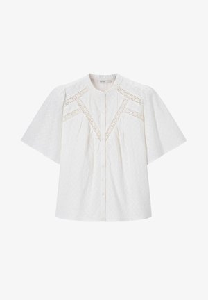 Jonge vrouw met lang bruin haar die een witte gestructureerde blouse met kanten details en lichtblauwe spijkerbroek draagt, staand met handen in haar zij.
