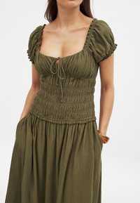 Olivengrünes Kleid mit kurzen Puffärmeln, einem quadratischen Ausschnitt, einem gerafften Oberteil und einem langen, fließenden Rock mit seitlichen Taschen.