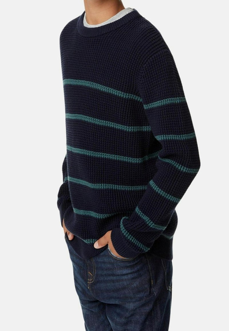 Personne portant un pull en maille bleu marine foncé avec des rayures horizontales turquoise, associé à un jean en denim bleu, mains dans les poches, sur un fond uni.