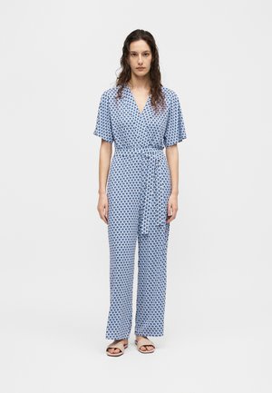 Kvinde med langt krøllet hår iført en blå og hvid jumpsuit med geometrisk mønster, korte ærmer og beige åbne sandaler.