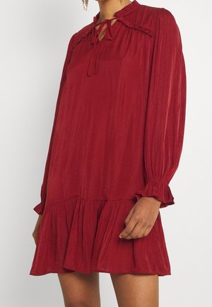 Woman wearing a loose-fitting, long-sleeve red dress with ruffled cuffs, ruffled collar, and tie detail at the neckline.