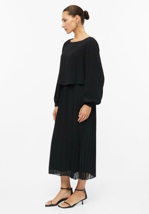 Femme en chemisier noir à manches longues et jupe midi plissée, portant des sandales noires à talons et des boucles d'oreilles dorées, debout de profil sur fond blanc.
