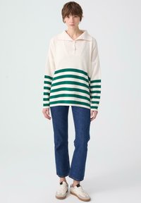 Witte pullover met groene horizontale strepen, geribbelde kraag en drie knopen; gecombineerd met blauwe rechte jeans en crèmekleurige sneakers.