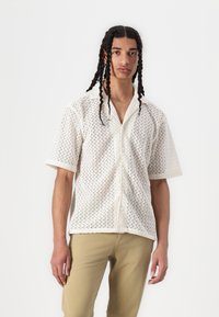 Chemise blanche à manches courtes avec un motif en zigzag et un col, dotée de fermetures à boutons et d'une coupe ample. Portée avec un pantalon kaki.