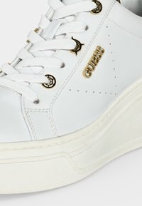 Λευκό sneaker GUESS με χρυσό λογότυπο και διακοσμητικές μεταλλικές οπές, λευκά κορδόνια και χοντρή υφή στην σόλα.
