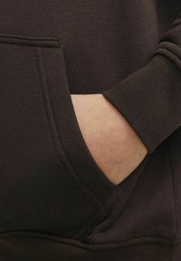 Tessuto di felpa marrone scuro con una consistenza morbida; presenta una tasca frontale e polsini a coste; mano inserita nella tasca.