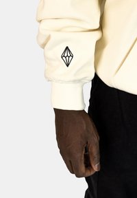 Crème sweatshirt met geribbelde manchetten, met een zwart geborduurd geometrisch logo op de linkermouw. Soepele textuur tegen donkere broeken.