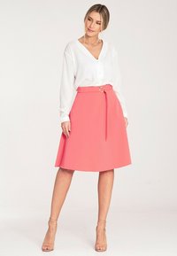 Koralowa spódnica w kształcie litery A z paskiem w talii, zakończona okrągłą metalową klamrą, połączona z białą bluzką z długim rękawem i beżowymi szpilkami.