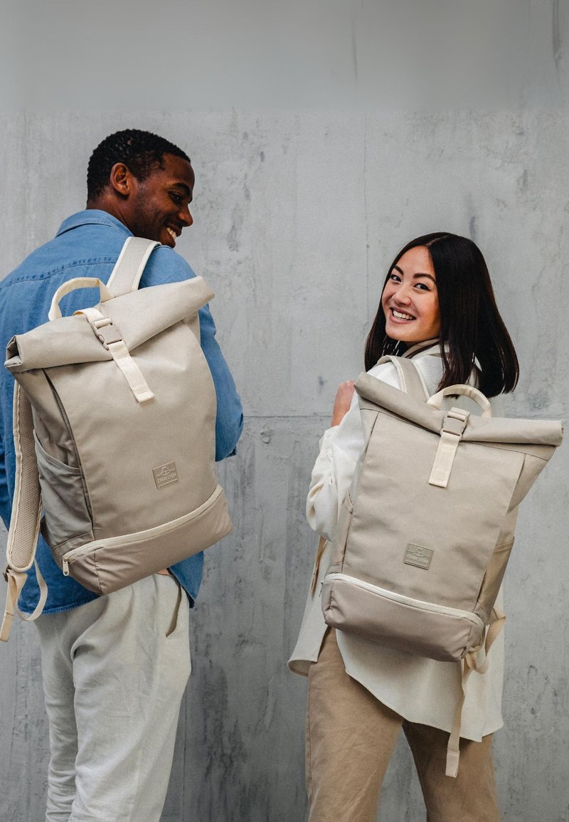 Két mosolygó ember bézs színű, felgombolt hátizsákkal áll egy szürke betonfal előtt, és hátranéznek a válluk felett.
